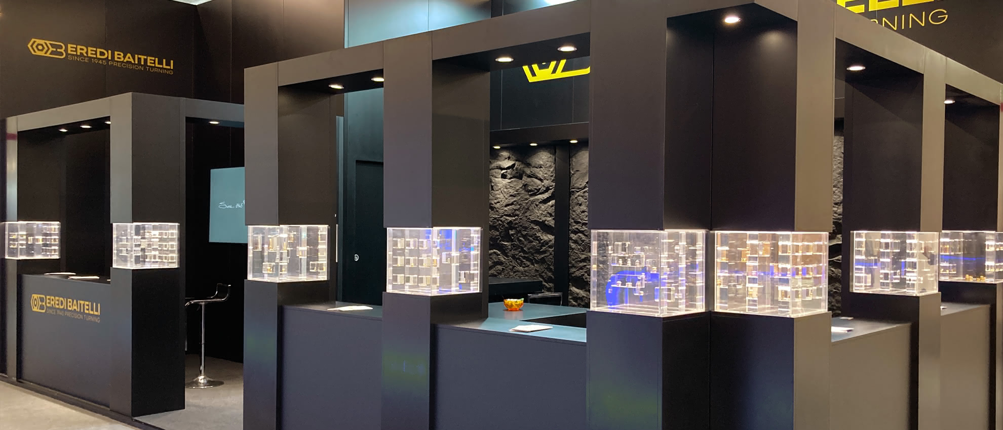 Vista laterale dello stand fieristico Eredi Baitelli, con elementi modulari neri e teche trasparenti disposte a isola.