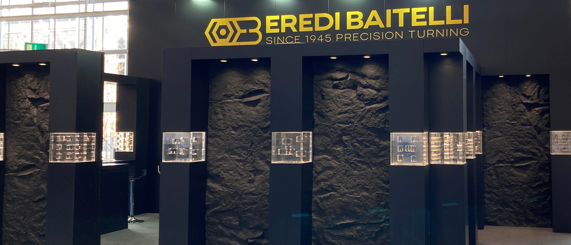 Vista frontale dello stand Eredi Baitelli, con logo aziendale e teche illuminate contenenti minuteria tornita di precisione.